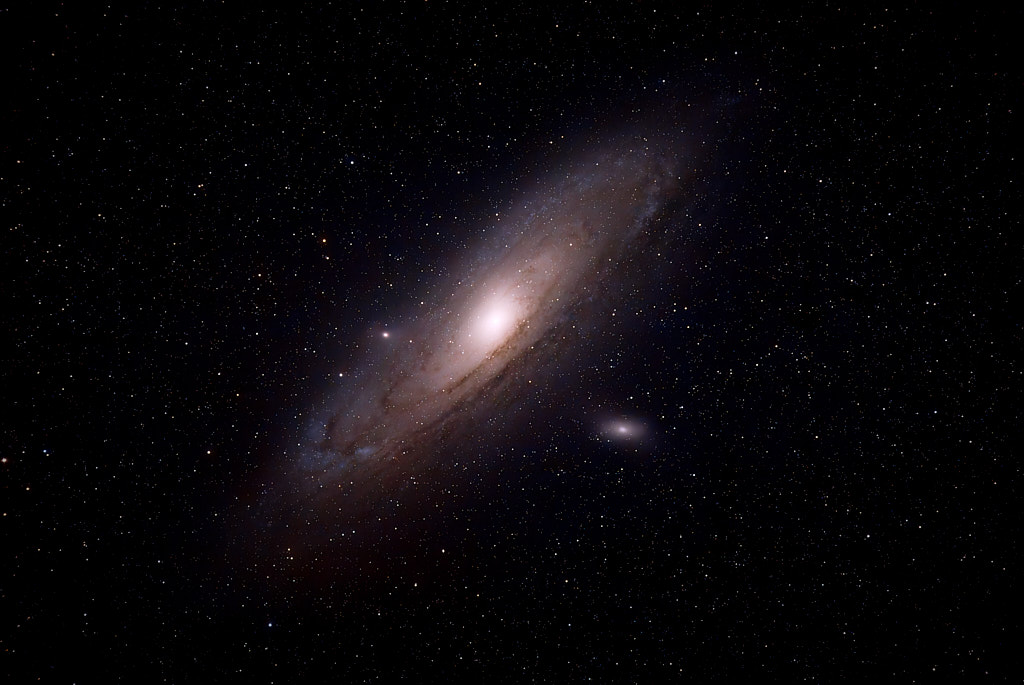 A large spiral galaxy, tilted 45 degrees in the frame, with a bright center, yellow and blue spiral arms with darker dust clouds around, and a small satellite galaxy below.