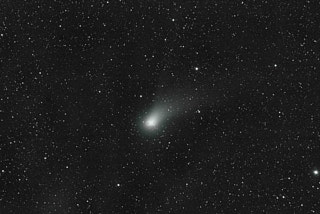A comet in the center of the frame, surrounded by a starfield, with its tail spreading out and trailing to the upper right corner.