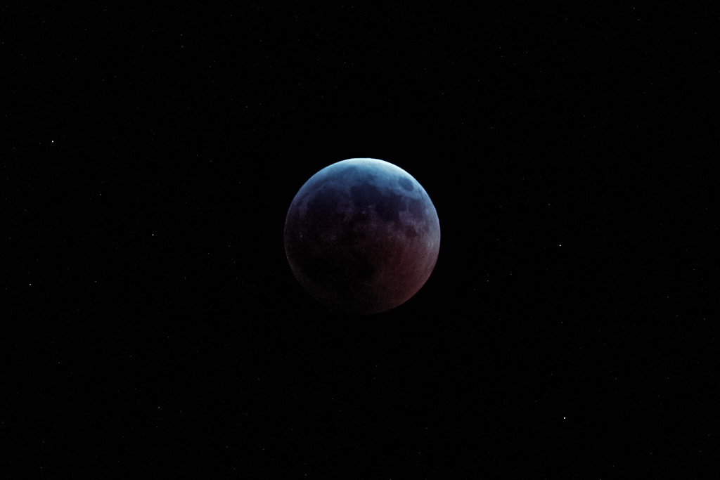 A not-quite eclipsed Moon, with its upper limb bright and the rest dark, with spreading red-coloring from the bottom right.