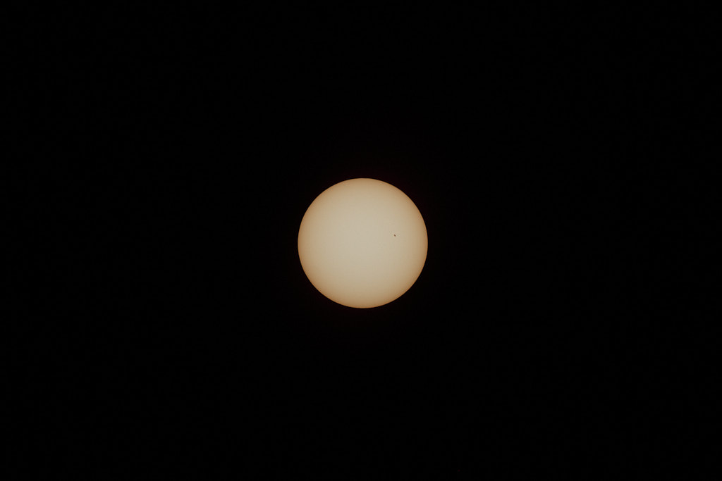 The sun, full and yellow, in the center of the frame, with a small, black sunspot on its upper right quarter.