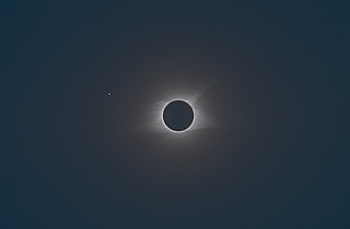 The sun obscured by the moon but for a tiny outline around it, with broad patterns in the corona visible. The star Regulus is seen to the lower left of the sun.