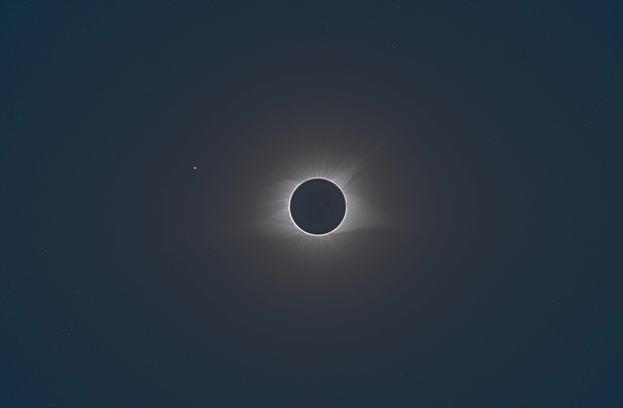 The sun obscured by the moon but for a tiny outline around it, with broad patterns in the corona visible. The star Regulus is seen to the lower left of the sun.
